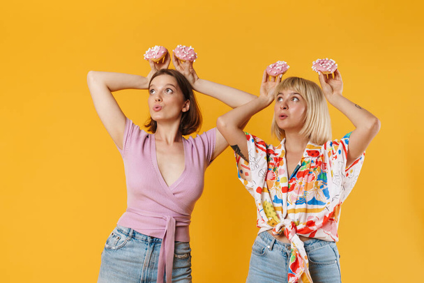 Portrait of two cheerful lovely girlfriends wearing summer clothes standing isolated over yellow background, showing tasty donuts, having fun - Foto, immagini