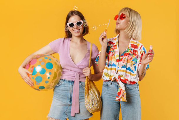 Portrait of two cheerful lovely girlfriends wearing summer clothes standing isolated over yellow background, holding beach ball, drinking beverage, blow soap bubbles - Foto, Imagem