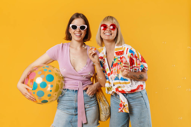 Portrait of two cheerful lovely girlfriends wearing summer clothes standing isolated over yellow background, holding beach ball, drinking beverage, blow soap bubbles - Foto, Imagem