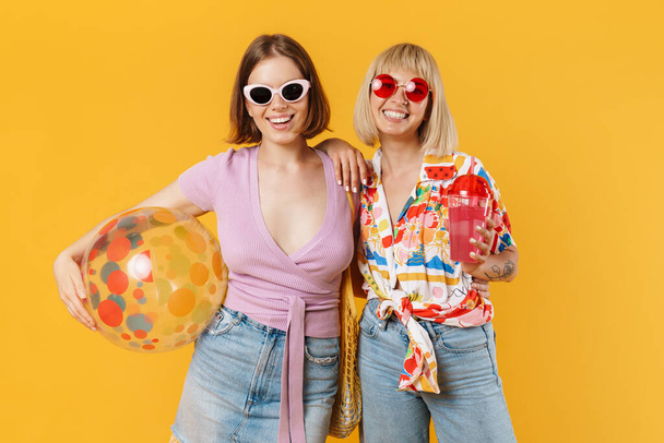 Portrait of two cheerful lovely girlfriends wearing summer clothes standing isolated over yellow background, holding beach ball, drinking beverage - Foto, Imagen