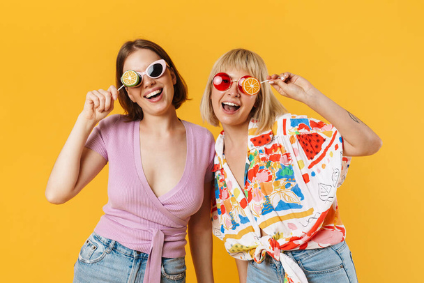 Portrait of two cheerful lovely girlfriends wearing summer clothes standing isolated over yellow background, holding lollipops - Photo, Image