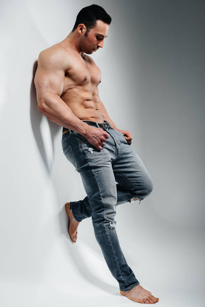 A young athlete bodybuilder poses in the studio topless in jeans near the wall. Sport. - 写真・画像