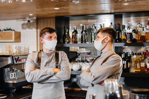 Za barem stojí dva styloví barmani v maskách a uniformách během pandemie. Práce restaurací a kaváren během pandemie. - Fotografie, Obrázek