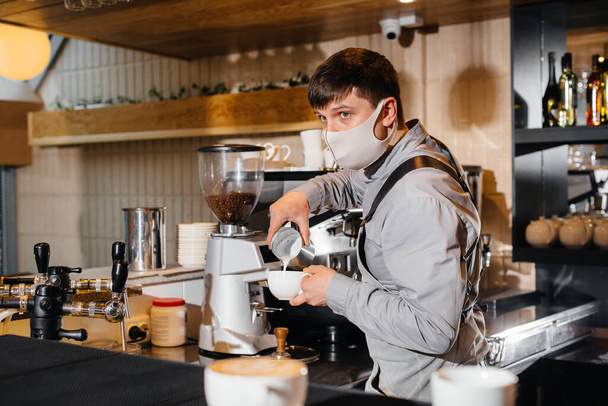 A masked barista prepares delicious coffee at the bar in a cafe. The work of restaurants and cafes during the pandemic. - 写真・画像