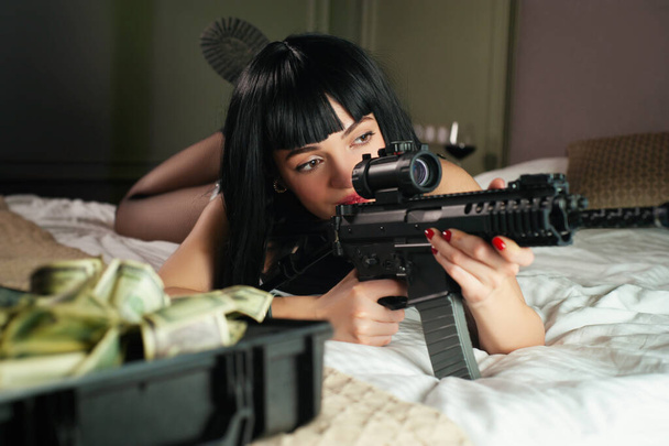 a female hitman in a hotel lies on a bed and holds an automatic rifle with a telescopic sight on the victim while committing murder - Photo, Image
