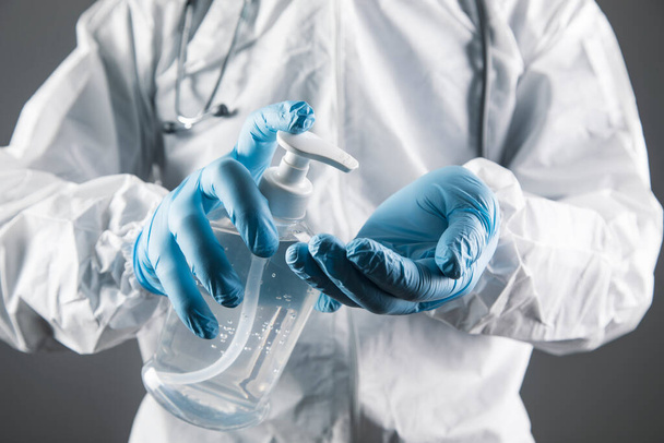 ένας γιατρός με λευκή προστατευτική στολή πλένει τα χέρια του με αντισηπτικό σε γκρι φόντο - Φωτογραφία, εικόνα