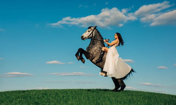 Bride in long white dress sits horseback on rearing horse against background of sky and clouds. With copy space. - Fotografie, Obrázek