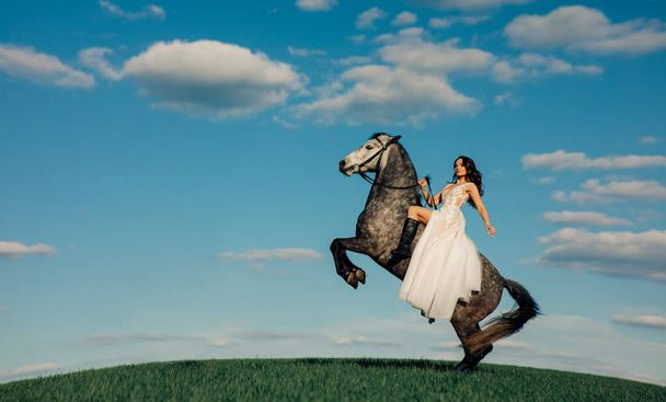 Bride in long white dress sits horseback on rearing horse against background of sky and clouds. With copy space. - 写真・画像