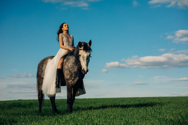 Bride in long white dress sits horseback on dapple grey horse on meadow against background of sky and clouds. - Fotografie, Obrázek