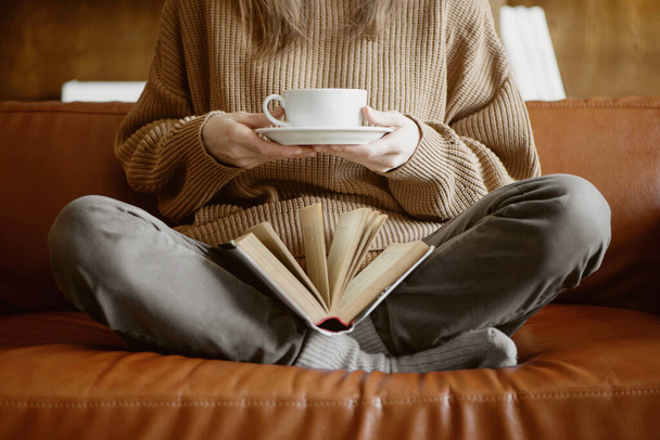 Cropped image of woman sitting on the couch holding cup of tea and reading a book. - Фото, изображение
