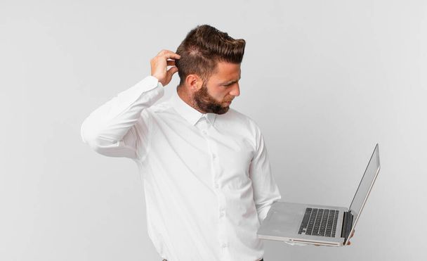 young handsome man feeling puzzled and confused, scratching head and holding a laptop - Fotoğraf, Görsel
