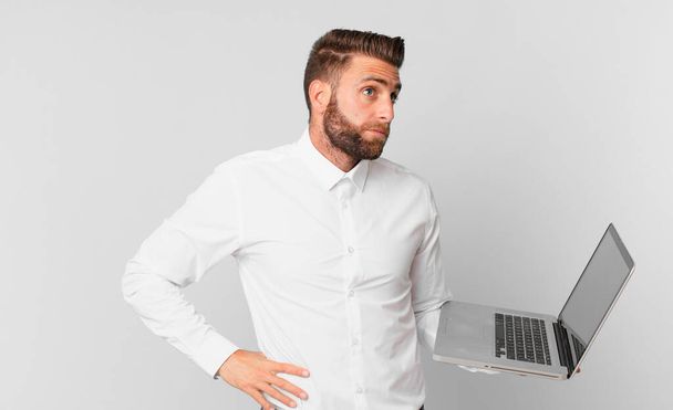 young handsome man shrugging, feeling confused and uncertain and holding a laptop - 写真・画像