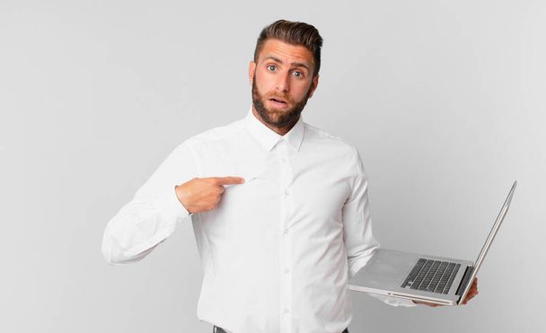 young handsome man looking shocked and surprised with mouth wide open, pointing to self and holding a laptop - Foto, Imagen