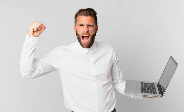 young handsome man shouting aggressively with an angry expression and holding a laptop - Foto, immagini