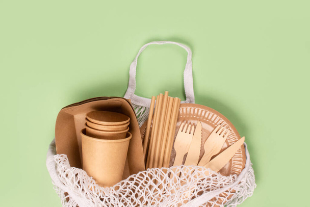 Flat lay, eco-friendly tableware, kraft paper food cups and containers with wooden cutlery in white cotton net bag on light green background with copy space. Street food take away paper packaging - Photo, Image
