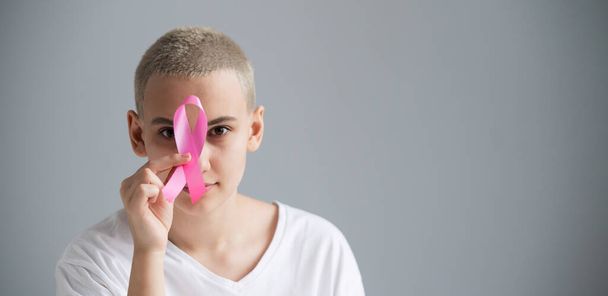 Young woman with short hair wearing a white t-shirt holding a pink ribbon as a symbol of breast cancer on a white background. - Photo, Image