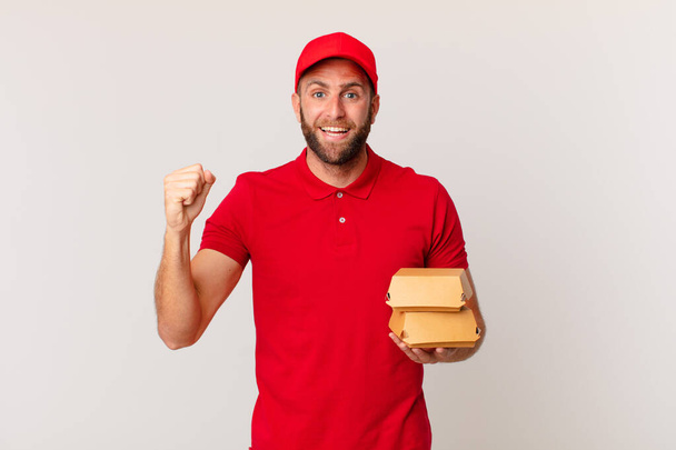 young handsome man feeling shocked,laughing and celebrating success burger delivering concept - Фото, изображение