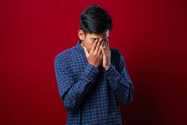 Ινδός με μουστάκι φοράει casual πουκάμισο με λυπητερή έκφραση καλύπτοντας το πρόσωπο με τα χέρια ενώ κλαίει. έννοια της κατάθλιψης σε κόκκινο φόντο - Φωτογραφία, εικόνα