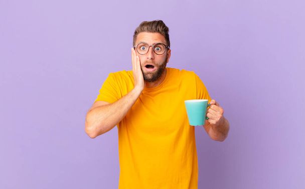 young handsome man feeling shocked and scared. and holding a coffee mug - Foto, Imagem