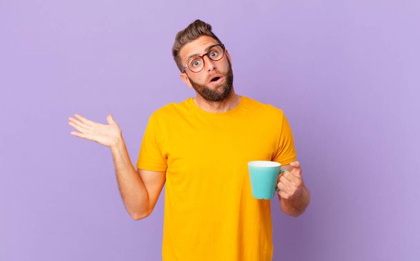 joven hombre guapo mirando sorprendido y sorprendido, con la mandíbula caída sosteniendo un objeto. y sosteniendo una taza de café - Foto, imagen