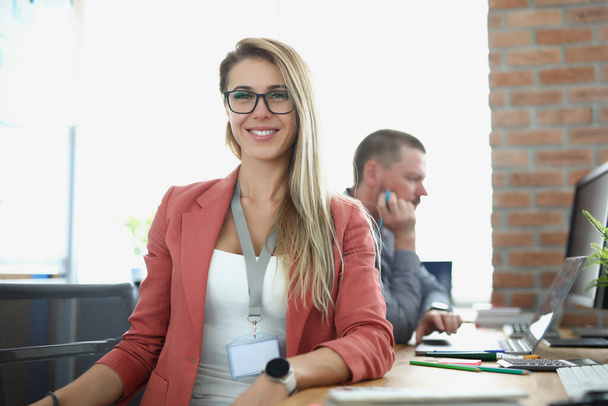 Молодая улыбающаяся деловая женщина сидит за рабочим столом на фоне делового офиса - Фото, изображение