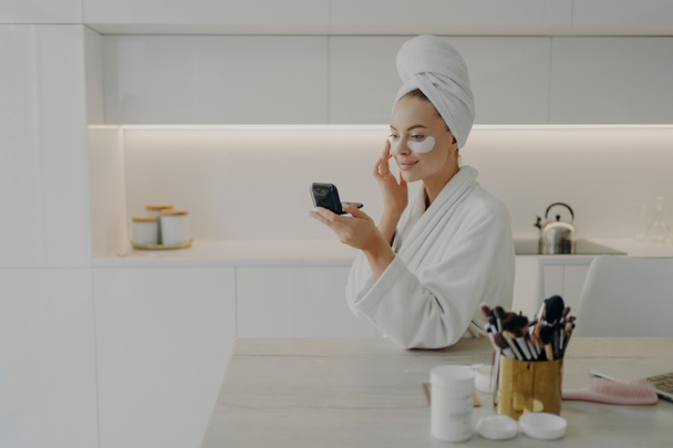 Mujer feliz en albornoz blanco con toalla en la cabeza aplicando parches cosméticos debajo de los ojos - Foto, Imagen