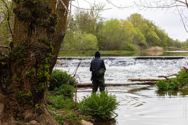 Young angler wearing a vest and high plastic boots, standing in the middle of the river to fish with a rod and tackle against the backdrop of a small waterfall in the photo selective focus - Photo, Image