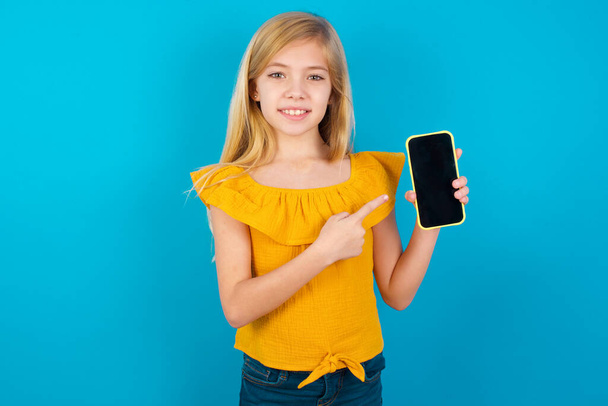 Улыбающаяся маленькая девочка, указывающая указательным пальцем на мобильный телефон с пустым экраном - Фото, изображение
