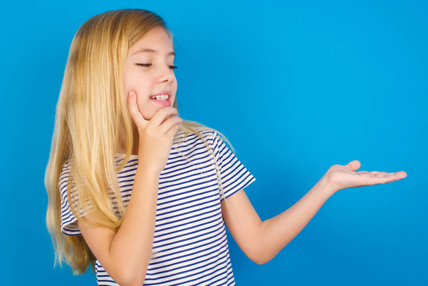 Funny Caucasian kid girl wearing striped shirt against blue wall holding open palm new product. I wanna buy it! - Фото, изображение
