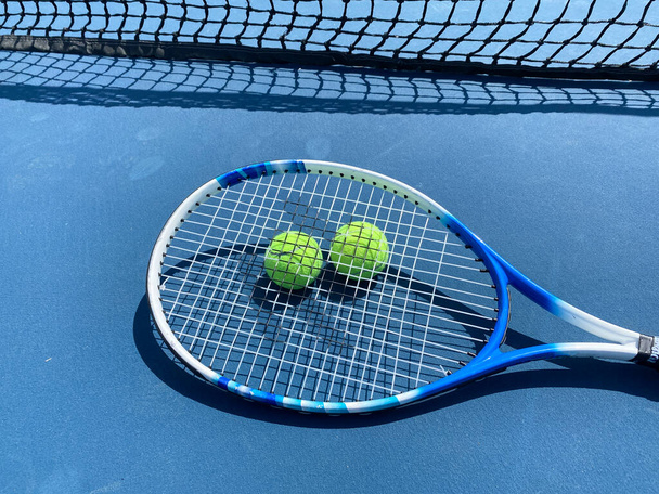 Μια ρακέτα του τένις ξαπλωμένη πάνω από δύο κίτρινες μπάλες του τένις σε ένα μπλε γήπεδο κοντά στο δίχτυ του τένις. - Φωτογραφία, εικόνα