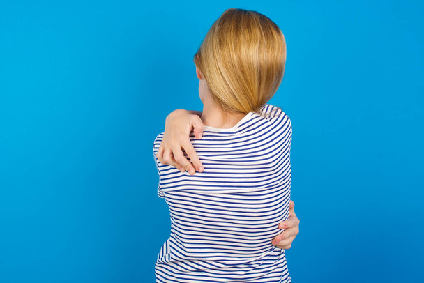 Caucasian girl wearing striped shirt against blue wall, hugging herself happy and positive from backwards. Self love and self care - Photo, Image