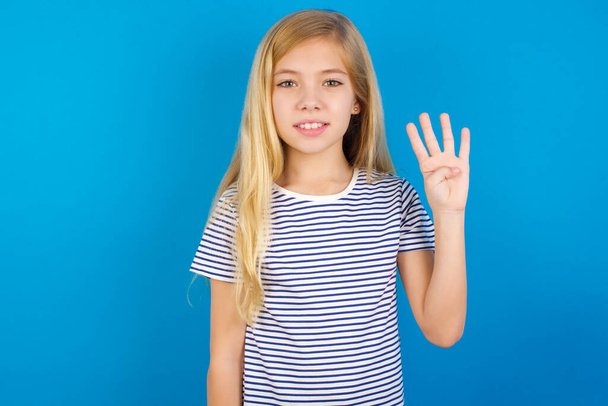 Chica caucásica con camisa a rayas contra la pared azul mostrando y apuntando hacia arriba con los dedos número cuatro mientras sonríe confiado y feliz. - Foto, Imagen