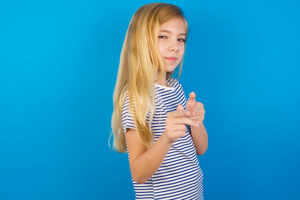 Fröhliches kaukasisches Mädchen trägt gestreiftes Hemd gegen blaue Wand, zwinkert und zeigt mit dem Zeigefinger in die Kamera, wählt jemanden aus, macht Fingerpistole - Foto, Bild