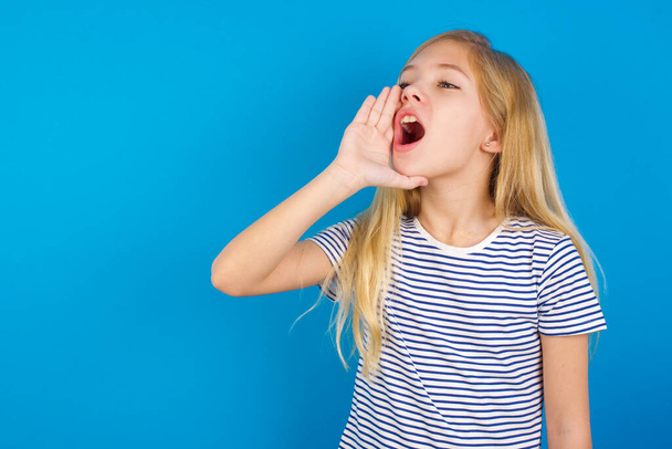 Caucasian girl wearing striped shirt against blue wall shouting and screaming loud to side with hand on mouth. Communication concept. - Foto, Bild