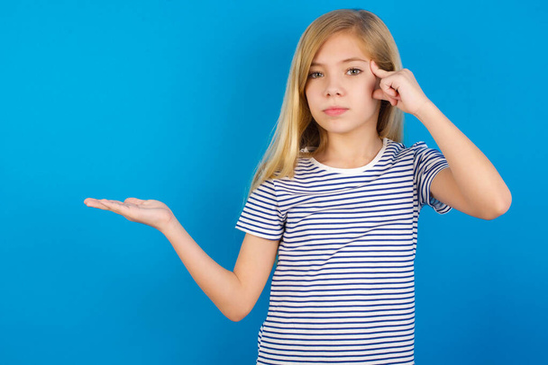Caucasian girl wearing striped shirt against blue wall confused and annoyed with open palm showing copy space and pointing finger to forehead. Think about it. - Photo, Image