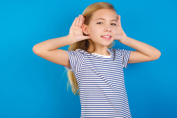 Белая девушка в полосатой рубашке на синей стене, пытается услышать обе руки на ушном жесте, любопытная для сплетен. Проблемы со слухом, глухой - Фото, изображение