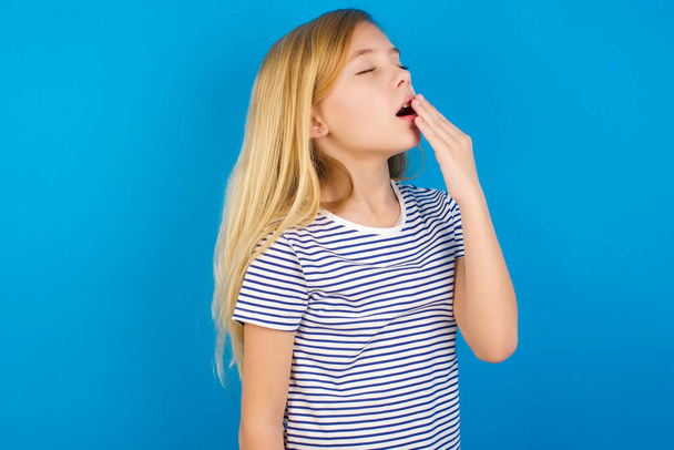 blank meisje dragen gestreept shirt tegen blauwe muur wordt moe en geeuwen  - Foto, afbeelding