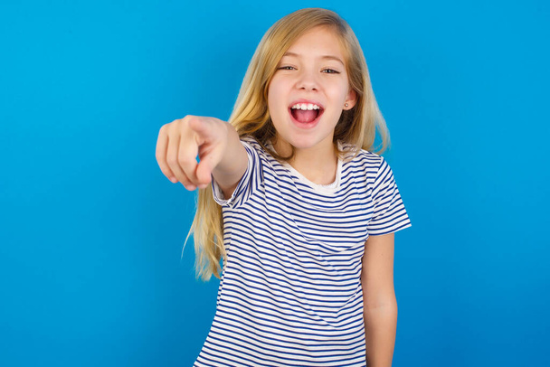 Menina caucasiana vestindo camisa listrada contra parede azul apontando descontente e frustrado para a câmera - Foto, Imagem