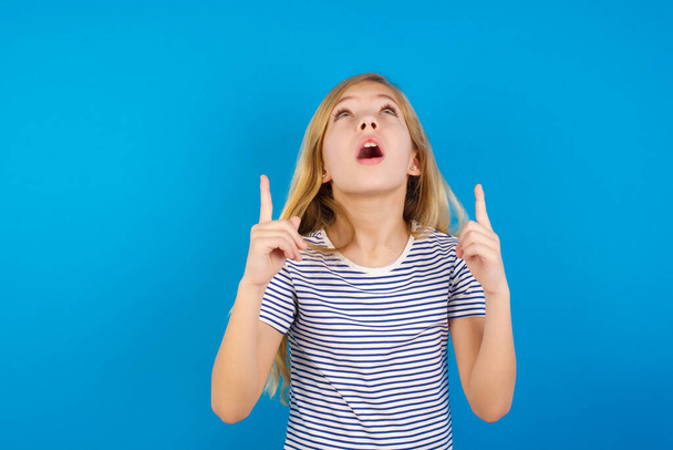 Kavkazská dívka v pruhované košili proti modré stěně užaslá a překvapená při pohledu nahoru a ukazuje prsty a zvednutými pažemi. - Fotografie, Obrázek