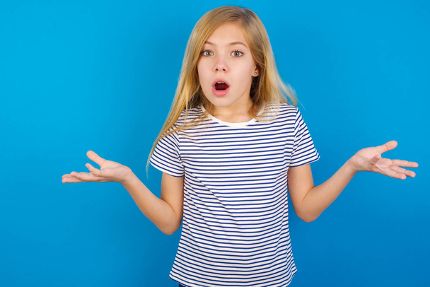Frustriertes kaukasisches Mädchen mit gestreiftem Hemd vor blauer Wand fühlt sich verwirrt und zögerlich, zuckt ratlos mit den Schultern, hält den Mund weit offen, weiß nicht, was zu tun ist. - Foto, Bild