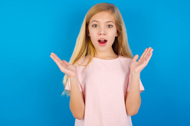 Surprised terrified Caucasian girl wearing pink T-shirt against blue wall Gestures with uncertainty, stares at camera, puzzled as doesn't know answer on tricky question. People, body language, emotions concept - Foto, afbeelding
