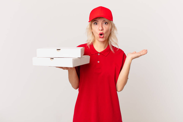 young pretty woman looking surprised and shocked, with jaw dropped holding an object. pizza delivering concept - Photo, Image