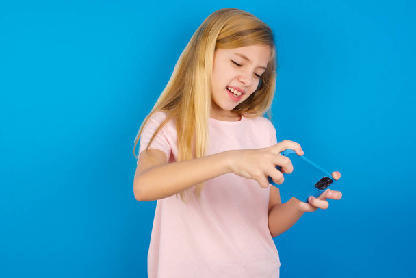 blank meisje dragen roze T-shirt tegen blauwe muur houden in handen cel spelen van video games of chatten - Foto, afbeelding
