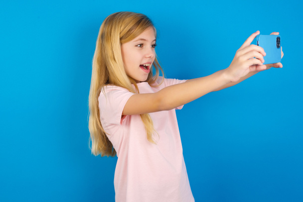 Niña caucásica usando camisa rosa contra la pared azul tomando una selfie para publicarla en las redes sociales o tener una videollamada con amigos. - Foto, Imagen