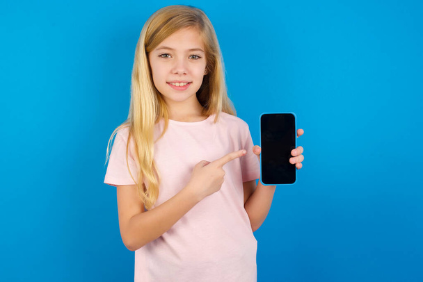 Hymyilevä valkoihoinen tyttö vaaleanpunaisessa paidassa sinistä seinää vasten. Osoittaminen etusormella matkapuhelimeen tyhjällä näytöllä - Valokuva, kuva