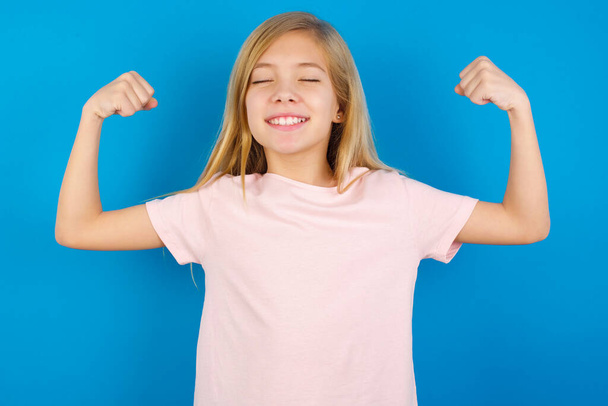 Sterke krachtige blanke meisje draagt roze shirt tegen blauwe muur tanden glimlach, heft armen en toont biceps. Kijk naar mijn spieren.! - Foto, afbeelding