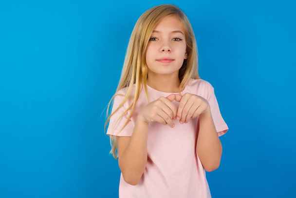 biały dzieciak dziewczyna noszenie różowy koszula przeciw niebieski ściana robi królicze łapki i spojrzenia z niewinny wyraz zabawy z jej mały dziecko - Zdjęcie, obraz