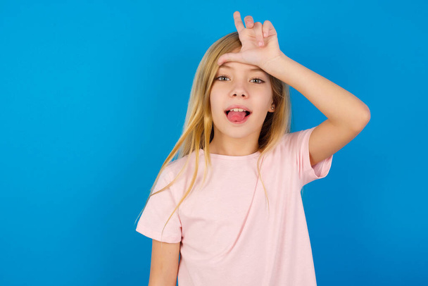 Caucasian kid girl wearing pink shirt against blue wall gestures with finger on forehead makes loser gesture makes fun of people shows tongue - Photo, Image