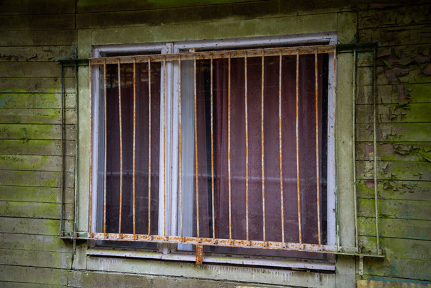 Окно старого деревянного дома с ржавыми металлическими решетками безопасности. Дом с шелушащейся краской из деревянных досок. Дом на дереве - Фото, изображение
