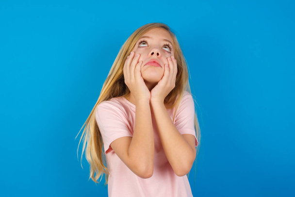 青い壁にピンクのシャツを着た白人の子供は頬に手を保つ不満そうな表情を退屈しています。ストレスのかかる絶望的なモデル - 写真・画像